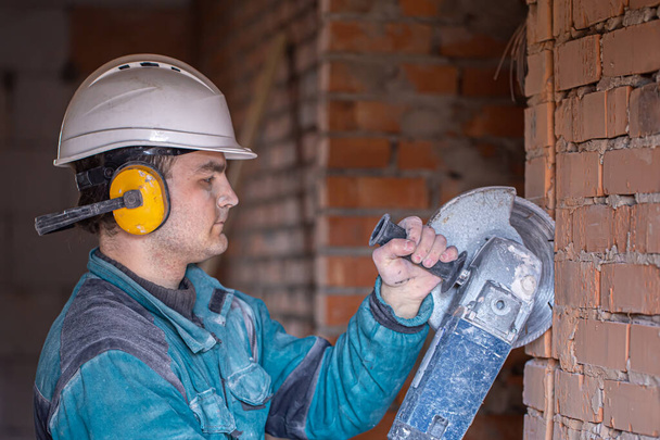 Primer plano de un constructor en un casco protector en una instalación de trabajo funciona con una herramienta de corte. - Foto, Imagen
