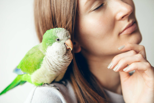 Zblízka sedí zelený papoušek a přitulí se k ženským ramenům. Roztomilý mnich Papoušek. Přírodní světlo, selektivní zaměření životní styl fotografie. Sdílení náklonnost s malým roztomilým ptákem - Fotografie, Obrázek