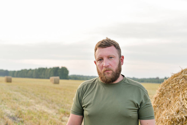 Un hombre con barba en el fondo de un pajar. Agricultor en un campo con heno - Foto, imagen