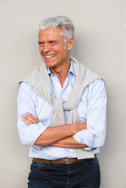 Portret van een glimlachende oudere man met armen gekruist door een witte muur die wegkijkt - Foto, afbeelding