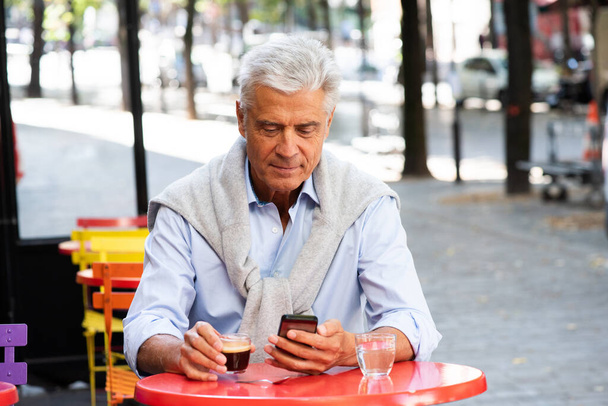 Ritratto di bell'uomo anziano seduto alla terrazza del caffè guardando il telefono cellulare  - Foto, immagini