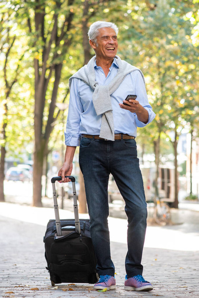 Retrato completo de un hombre mayor sonriente parado en la ciudad con teléfono celular y maleta - Foto, imagen