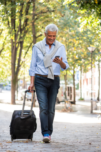 Full body portrait of older man walking with mobile phone and suitcase on city street - Foto, Imagen
