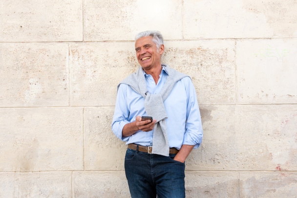 Εμπρός πορτρέτο του χαρούμενος ηλικιωμένος άνδρας ακουμπά στον τοίχο με κινητό τηλέφωνο στο χέρι - Φωτογραφία, εικόνα