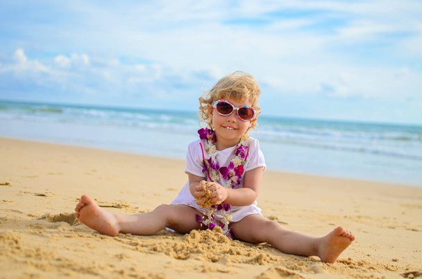 Una niña blanca de tres años con una cuenta de flores juega en la arena de la playa. - Foto, Imagen