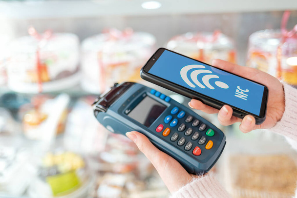 A woman's hands is holding a payment terminal and paying for a purchase using a smartphone.On the phone screen, the wi-fi network.Showcase with cakes in the background. The concept of NFC, business and banking transactions . - Photo, Image