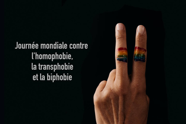 de regenboogvlag in de vingers van een jonge persoon die het V-teken doet en de tekst internationale dag tegen homofobie, fobie en bifobie geschreven in het Frans tegen een zwarte achtergrond - Foto, afbeelding
