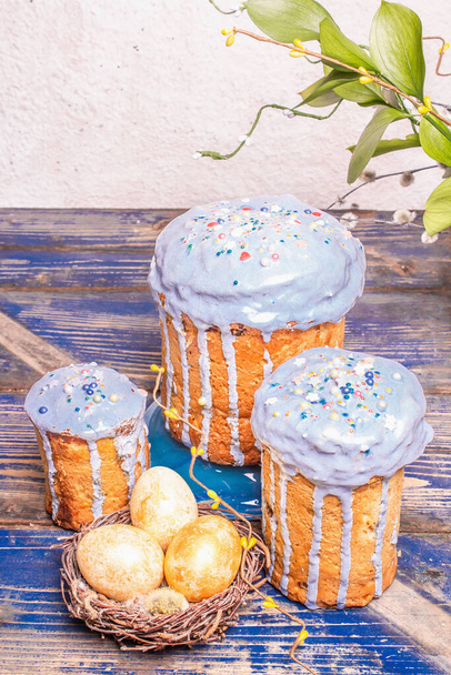 Easter cake with blue icing, topping, edible beads. Natural healthy ingredients, golden eggs in a nest, festive decor. Modern stands, old wooden table background, copy space - Photo, Image