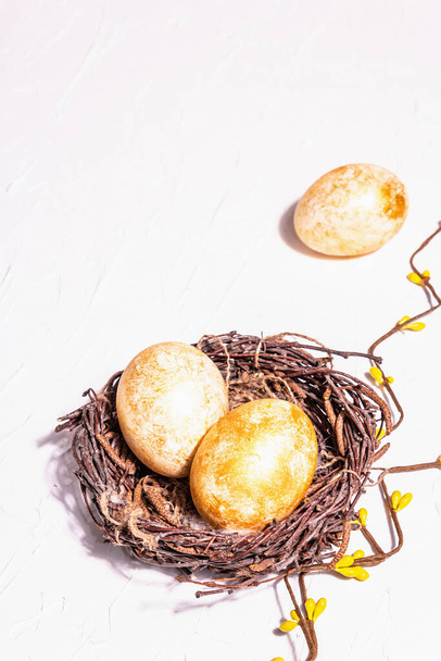 Uova di Pasqua d'oro in un nido di vimini. Simbolo tradizionale pasquale, arredamento festivo. Trendy luce dura, ombra scura, sfondo in gesso bianco, posto per il testo - Foto, immagini