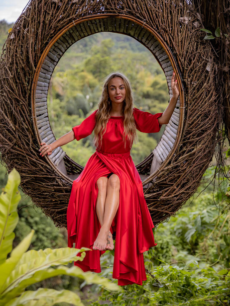 Bali trend. Straw nest in tropical forest. Caucasian woman in long red dress taking photo in a straw nest. Vacation in Asia. Travel lifestyle. Summer concept. Bongkasa, Bali, Indonesia - Fotó, kép