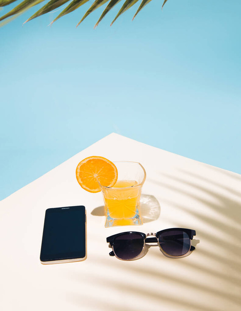 Composition tropicale estivale avec du jus d'orange jaune dans un verre. Ombre des feuilles de palmier. Lunettes de soleil et téléphone sur la table. - Photo, image