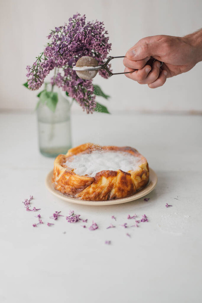 Sprinkling icing sugar over freshly baked ricotta cheesecake on white table with lilac flower in glass vase. Homemade rustic cake with organic ingredients. Flourless pie. Food styling, selective focus - Photo, Image