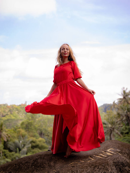 Bali trendfotó. Fehér nő hosszú piros ruhában, nagy kövön áll a trópusi esőerdőben. Ázsiai vakáció. Utazási életmód. Nyári koncepció. Lélegzetelállító kilátás. Bongkasa, Bali, Indonézia - Fotó, kép