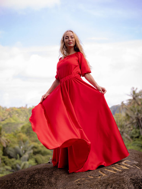 バリ島のトレンド写真。熱帯雨林の大きな石の上に立って長い赤いドレスを着た白人女性。アジアでの休暇。旅行生活。夏のコンセプト。息をのむような景色。インドネシアのバリ島、ボンカサ - 写真・画像