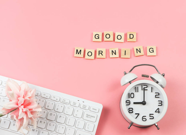 Top view or flat lay of white vintage alarm clock  with bouquet of pink flower on computer keyboard and wooden letter GOOD MORNING  on pink background. - Photo, Image