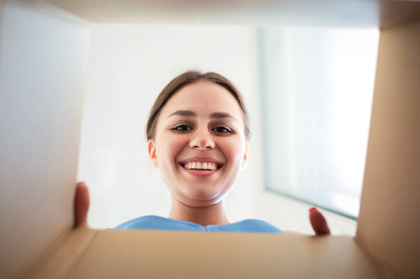 Знизу щаслива молода жінка посміхається і дивиться всередину картонної коробки, відкриваючи замовлені товари
 - Фото, зображення