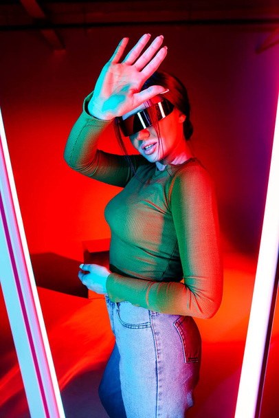 Young female in futuristic glasses gesturing stop and protecting face from bright neon light in modern room - Photo, Image