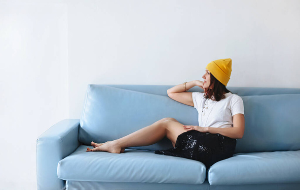 Widok z boku boso kobieta w modne ubrania patrząc na aparat podczas siedzenia na niebieskiej kanapie na szarym tle - Zdjęcie, obraz
