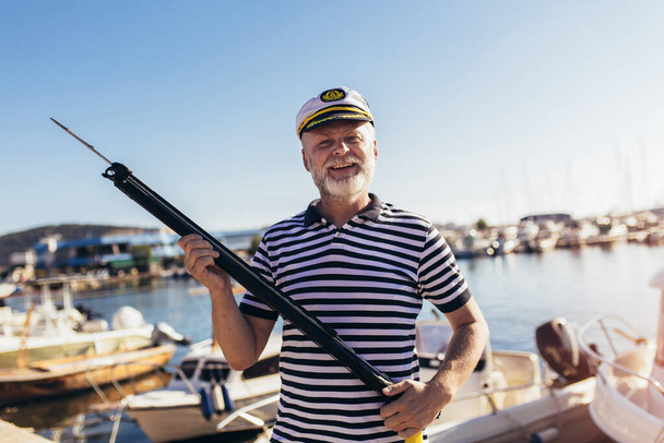 Starší muž stojící u moře oblečený v námořnické košili a klobouku s harpunou - Fotografie, Obrázek