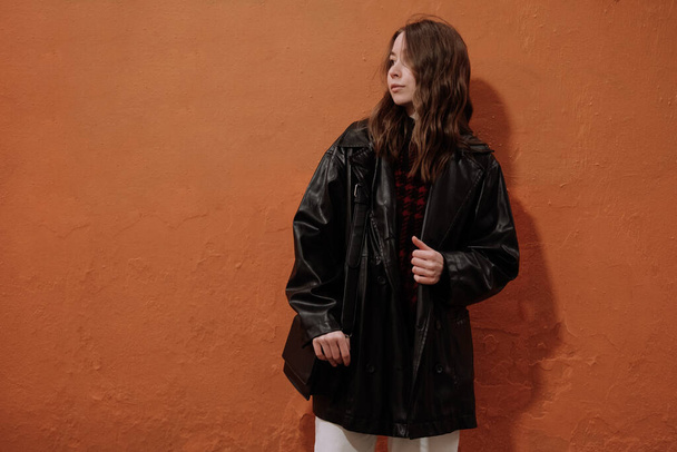 Улыбающаяся молодая женщина в черной кожаной куртке, стоящая перед оранжевой стеной, глядя налево, копирует пространство. Городской стиль и уличная мода. Девушка с кудрявой прической в повседневной одежде - Фото, изображение