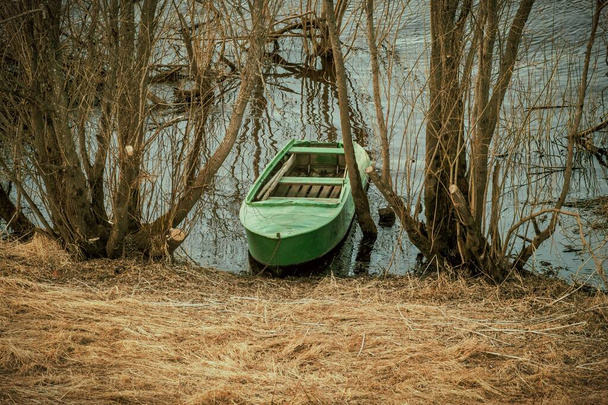 Antiguo barco de metal verde entre los árboles en la orilla del río. Primavera paisaje retro - Foto, imagen