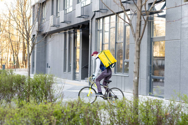 Young courier delivering food with a yellow thermal backpack, riding a bicycle in the city. Food delivery service concept - Fotó, kép