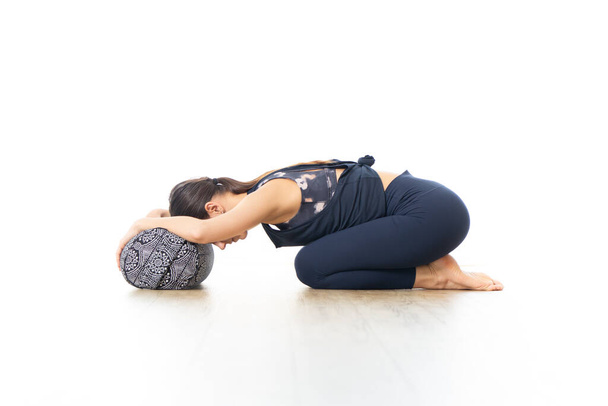 Restorative yoga with a bolster. Young sporty attractive woman in bright white yoga studio, lying on bolster cushion, stretching and relaxing during restorative yoga. Healthy active lifestyle - Photo, Image