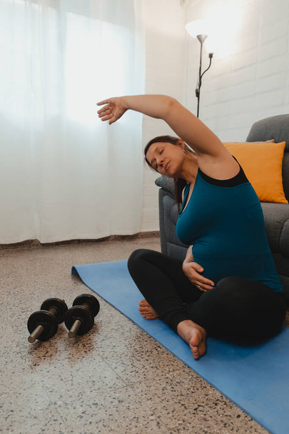 Vertical shot of a pregnant Latina woman stretching after yoga and exercise. Concept of healthy life during pregnancy and exercise at home. - Photo, Image