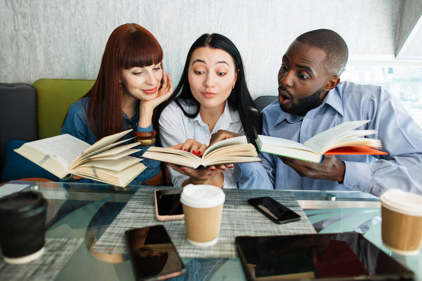 Book lovers, students, self-education concept. Three surprised excited multiethnic friends, sitting at the table in cafe and reading books, looking and pointing at interesting site. - Photo, Image