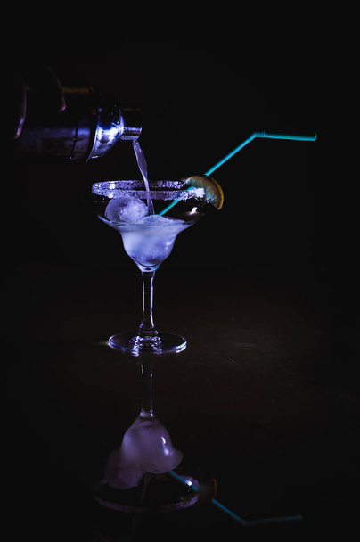 Охлажденный коктейль "Маргарита", смешанный с текиллой Куантро и лимонным соком, выливаемым из коктейльного шейкера на кубики льда с солью на краю бокала и украшенным лимоном и голубой соломой в рождественскую ночь - Фото, изображение