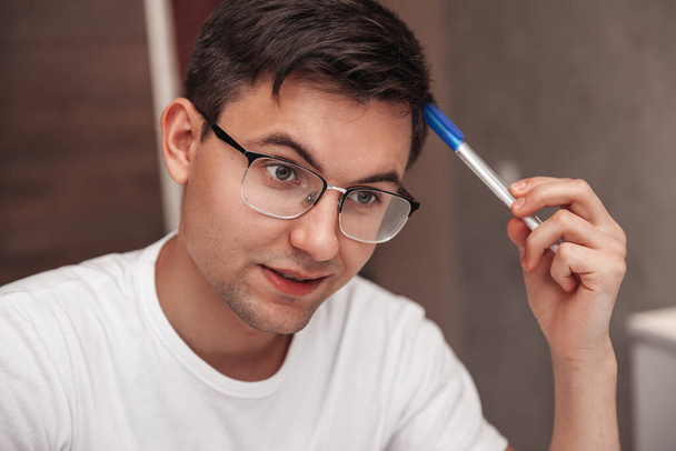 A pensive young man rubs his head with a ballpoint pen. Close-up portrait - Zdjęcie, obraz