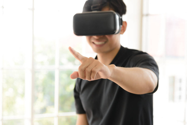 VR仮想現実ヘッドセットや自宅のリビングルームで指で指差す3Dメガネで経験を積んだ若いアジア人男性。テクノロジー、ゲーム、エンターテイメントのコンセプト - 写真・画像
