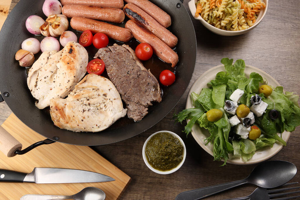 Mix meet chicken beef steak sausage tomato onion garlic fry grill oil metal fry pan colourful pasta green salad with cheese olive mint basil pesto sauce board knife fork spoon rustic wooden table - Foto, imagen