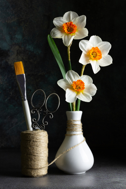 白い花瓶、装飾はさみ、ツインと暗い背景にブラシをペイントで水仙やナルシスの美しい花束。創造的な静止生活の概念。スペースのコピー. - 写真・画像