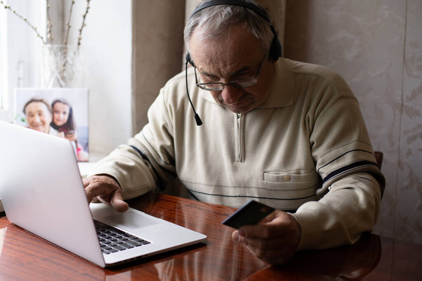 Un homme âgé souriant porte des écouteurs d'onde à la caméra ayant un appel vidéo sur un ordinateur portable, un homme âgé heureux dans un casque assis sur un canapé à la maison parle en utilisant des technologies modernes et une connexion sans fil - Photo, image