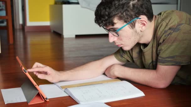 Ein netter kaukasischer Junge macht seine Hausaufgaben und studiert auf dem Parkett in seinem Zimmer. Er bedient sich am Tablet, in der Nähe der Blätter, auf denen er auch das Smartphone schreibt.. - Filmmaterial, Video