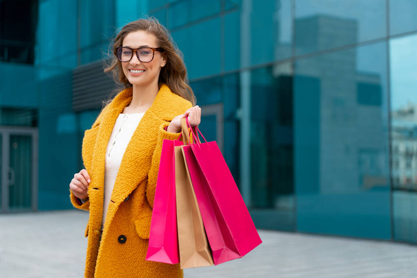 Business woman with shopping bags dressed yellow coat walking outdoors corporative building background Caucasian female business person near office building Stylish businesswoman season sale - Foto, afbeelding