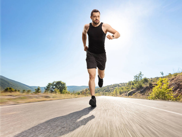 Full length portrait of a fit man with a smartwatch running on an open asphalt road - Фото, зображення