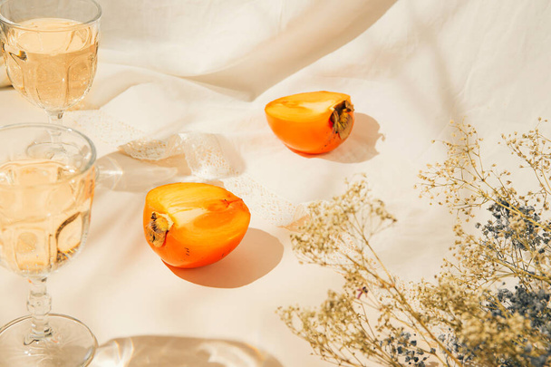 Halfs of persimmon fruits and two glasses with lemonade on pastel background with white cloth and sunlit. Summer drinks and refreshment concept. Minimal style composition. - Photo, Image