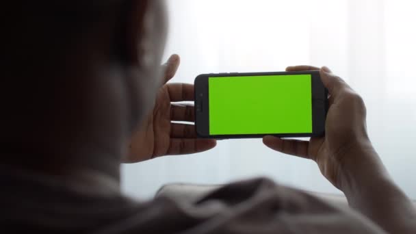 Hombre afroamericano usando smartphone con pantalla verde, viendo películas, teléfono con croma key en posición horizontal - Metraje, vídeo