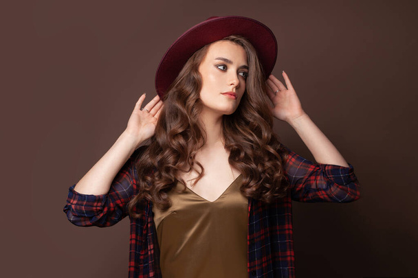 Elegante donna in camicia a quadri e cappello fedora in posa su sfondo marrone - Foto, immagini