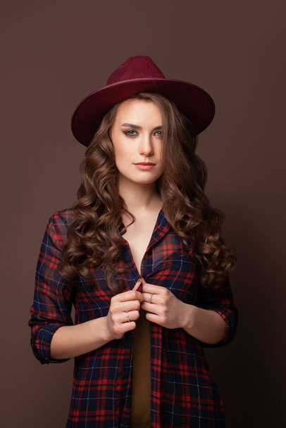 Brunette woman in plaid shirt and fedora hat on brown background - Foto, Imagen