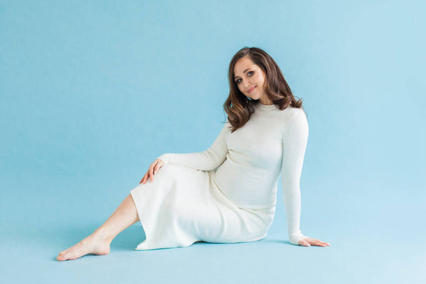 Ευτυχισμένη εγκυμοσύνη. Χαριτωμένη έγκυος γυναίκα κάθεται σε μπλε φόντο - Φωτογραφία, εικόνα
