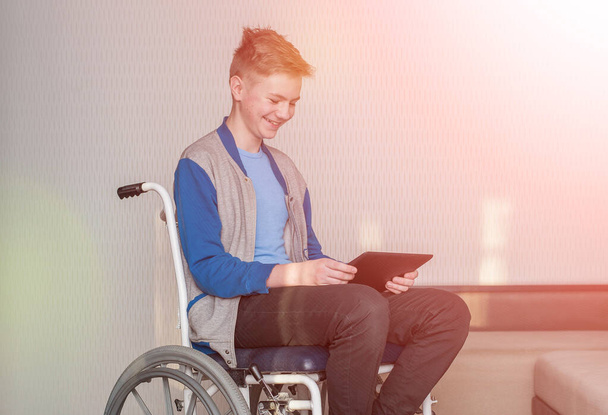 έφηβος μαθητής αγόρι κάθεται σε μια αναπηρική καρέκλα, ενώ χρησιμοποιώντας ένα tablet στο σπίτι. - Φωτογραφία, εικόνα