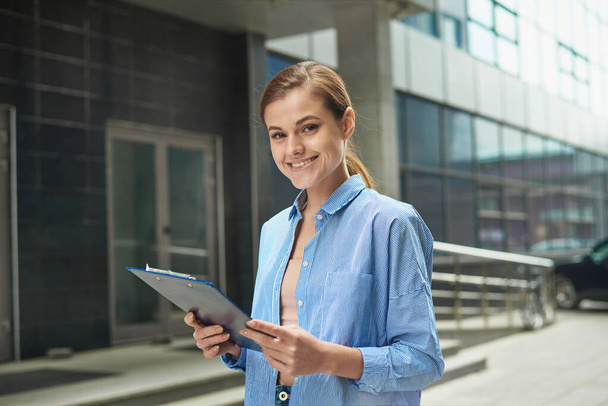 Jonge vrouwelijke zakenvrouw die een tablet vasthoudt met documenten in haar handen en glimlachend naar de camera kijkt. Het concept van werken in het kantoor van jonge professionals - Foto, afbeelding