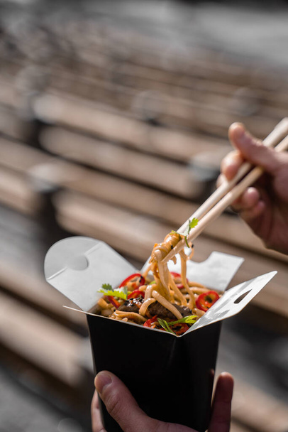 Wok σε κουτί σε μαύρο δοχείο τροφίμων. Κρατώντας πικάντικα νουντλς με ξυλάκια. Υπηρεσία παράδοσης γρήγορου φαγητού. Χοιρινό κινέζικο γεύμα στο δρόμο - Φωτογραφία, εικόνα
