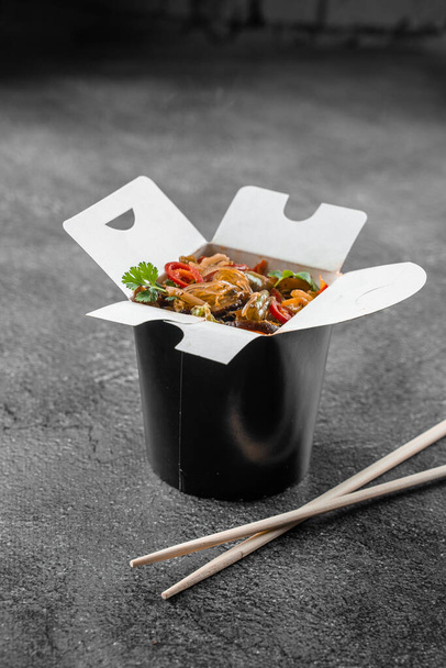 黒い食品容器の中のボックス米麺で中華鍋.ファーストフードの配達サービス。テイクアウト中華街の食事 - 写真・画像