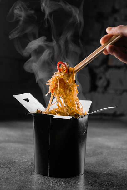 Kouřící rýžové nudle ve wok boxu na černém pozadí. Služba rychlého občerstvení. Jídlo z čínských ulic. Udon v černé nádobě na jídlo - Fotografie, Obrázek