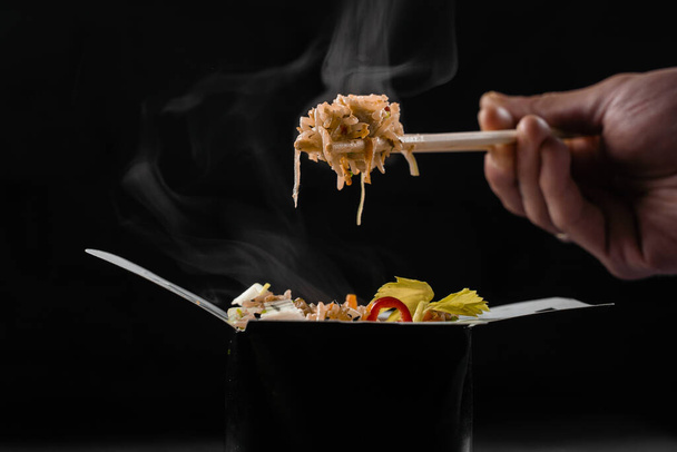 Arroz caliente al vapor en caja wok sobre fondo negro. Sostiene arroz con palillos chinos. Plato picante asiático para restaurante callejero y servicio de entrega de comida rápida - Foto, imagen