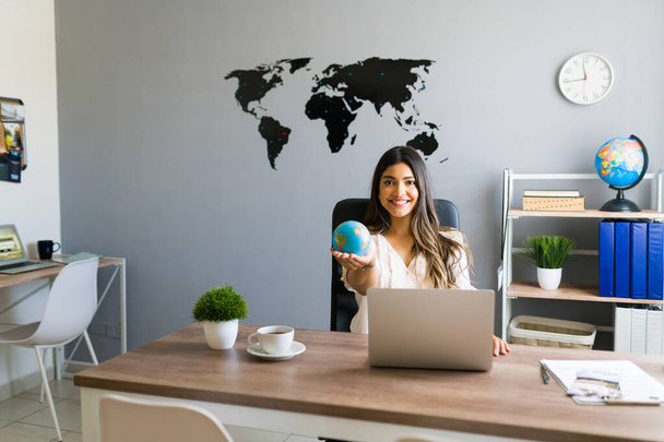 De wereld in mijn handen houden. Aantrekkelijke vrouwelijke reisagent met een globe en glimlach tijdens het werken in haar moderne kantoor  - Foto, afbeelding
